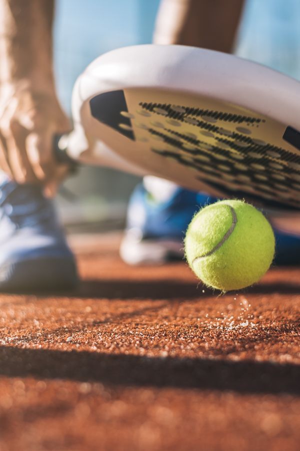 A close-up of a person holding a pickleball paddle near the ground as a green pickleball bounces on a brown court surface.